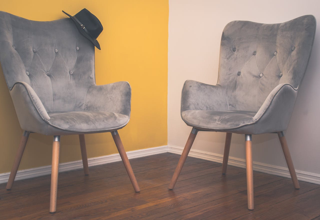 hero-img Two contemporary gray armchairs in a minimalist interior with a yellow accent wall and wooden flooring.